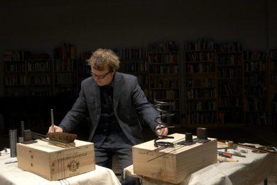 Ross Karre, a musician wearing a grey suit, stands behind two tables of various items such as springs, brushes, and rods on two wooden boxes.